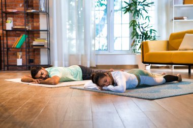 Indian Asian young couple practicing Makarasana or crocodile resting pose yoga together in their lavish home, focusing on relaxation and wellness clipart