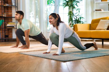 Indian Asian young couple joyfully practicing yoga together in their lavish home, executing Ek Pad Prasarita Padasana, or One-Legged Wide-Legged Forward Bend pose clipart