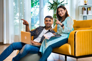 Hintli Asyalı genç bir çift gösterişli bir evde sarı bir kanepeye oturmuş, mali durumlarını birlikte idare ederken gülümsüyorlardı, bir dizüstü bilgisayar ve belgeleri bütçe ve gider planlaması için kullanıyorlardı.