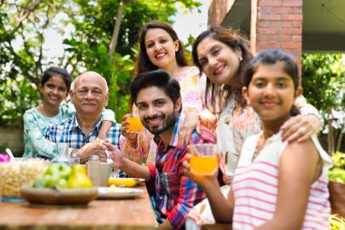 Altı kuşaklı Hintli Asyalı aile kahvaltı masasının etrafında poz veriyor çimlerin üzerinde, sabah ışığında mutlu anların tadını çıkarıyorlar.