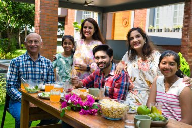 Altı kuşaklı Hintli Asyalı aile kahvaltı masasının etrafında poz veriyor çimlerin üzerinde, sabah ışığında mutlu anların tadını çıkarıyorlar.