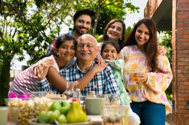 Altı kuşaklı Hintli Asyalı aile kahvaltı masasının etrafında poz veriyor çimlerin üzerinde, sabah ışığında mutlu anların tadını çıkarıyorlar.