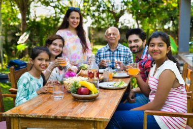 Altı kuşaklı Hintli Asyalı aile kahvaltı masasının etrafında poz veriyor çimlerin üzerinde, sabah ışığında mutlu anların tadını çıkarıyorlar.