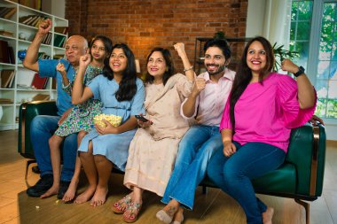 Hintli Asyalı altı nesillik aile evdeki bir kanepede birlikte oturur, heyecanlı bir şekilde televizyon izler, patlamış mısır yer ve uzaktan kumandayı tutarlar.