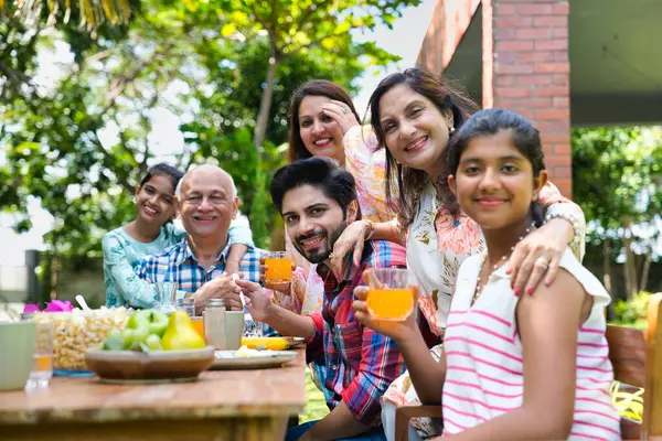 Altı kuşaklı Hintli Asyalı aile kahvaltı masasının etrafında poz veriyor çimlerin üzerinde, sabah ışığında mutlu anların tadını çıkarıyorlar.