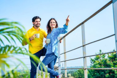 Genç Hintli çift mavi gökyüzünün altındaki teras balkonunda sabah kahvesinin keyfini çıkarıyor. Sıcak güneş ışığında rahat bir sohbet ediyorlar.