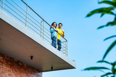 Genç Hintli çift mavi gökyüzünün altındaki teras balkonunda sabah kahvesinin keyfini çıkarıyor. Sıcak güneş ışığında rahat bir sohbet ediyorlar.