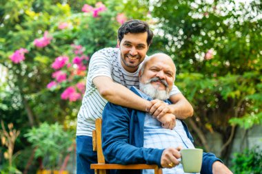 Hintli Asyalı mutlu yaşlı baba ev bahçesinde sandalyede oturuyor, genç oğul ona arkadan sarılıyor, kameraya gülümsüyor, baba-oğul bağını ve sevgisini kutluyor.