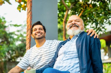 Hintli Asyalı mutlu yaşlı baba ve genç yetişkin oğul açık hava evinin verandasında oturuyor, Yürekten bir sohbete girişiyor ve birlikte gülüyorlar, Baba-oğul bağlarını gösteriyorlar