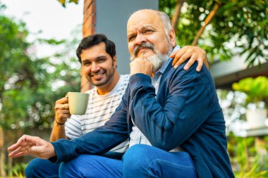 Hintli Asyalı mutlu yaşlı baba ve genç yetişkin oğul açık hava evinin verandasında oturuyor, Yürekten bir sohbete girişiyor ve birlikte gülüyorlar, Baba-oğul bağlarını gösteriyorlar