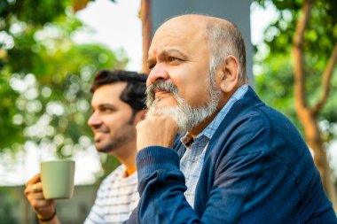 Hintli Asyalı mutlu yaşlı baba ve genç yetişkin oğul açık hava evinin verandasında oturuyor, Yürekten bir sohbete girişiyor ve birlikte gülüyorlar, Baba-oğul bağlarını gösteriyorlar