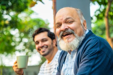 Hintli Asyalı mutlu yaşlı baba ve genç yetişkin oğul açık hava evinin verandasında oturuyor, Yürekten bir sohbete girişiyor ve birlikte gülüyorlar, Baba-oğul bağlarını gösteriyorlar