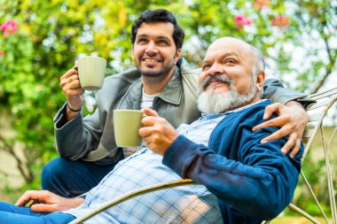 Closeup of Indian Asian Old Aged Father Relaxing on Chair in Home Garden as Young Adult Son Joins with Coffee Mug, Sharing Smiles and a Heartfelt Conversation clipart