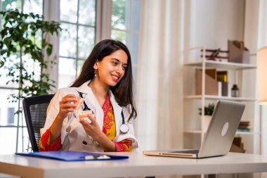 Hintli Asyalı genç dişçi beyaz önlüklü, dizüstü bilgisayarlı bir masada oturuyor, video görüşmesi sırasında takma diş kullanarak oral sağlık hizmetlerini anlatıyor.