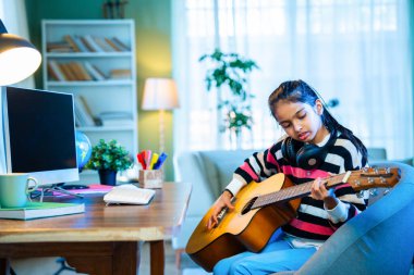 Hintli küçük kız evde online video dersinden akustik gitar çalmayı özenle öğreniyor, sanal müzik dersleri için bir masaüstü bilgisayarı kullanıyor, dijital öğrenme yoluyla yeteneklerini geliştiriyor.