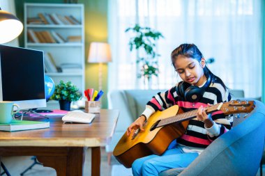 Hintli küçük kız evde online video dersinden akustik gitar çalmayı özenle öğreniyor, sanal müzik dersleri için bir masaüstü bilgisayarı kullanıyor, dijital öğrenme yoluyla yeteneklerini geliştiriyor.