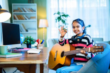 Hintli küçük kız evde online video dersinden akustik gitar çalmayı özenle öğreniyor, sanal müzik dersleri için bir masaüstü bilgisayarı kullanıyor, dijital öğrenme yoluyla yeteneklerini geliştiriyor.