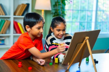 Hindistanlı şirin küçük kız ve erkek kardeşler tuvalde oyun oynuyorlar evde küçük bir adada fırça kullanıyorlar, yaratıcılığı ve sanat becerilerini araştırıyorlar modern evde masada birlikte oturuyorlar.