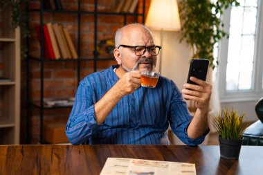 Hintli emekli adam internet okumak veya internette gezinmek için akıllı telefon kullanıyor. Ev masasında sade kahveyi yudumlarken, rahat görünüyor ve güncel konuları güncel tutuyor.