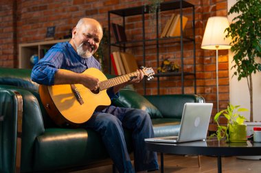 Kurta sakallı Hintli emekli bir adam, modern bir oturma odasındaki bir koltukta oturuyor, bilgisayarındaki online bir dersten akustik gitar çalmayı öğreniyor, müzik çalışmasının keyfini çıkarıyor.