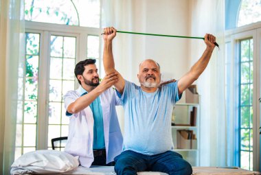 Hintli Asyalı fizyoterapist, direnç bandı egzersizleri yoluyla omuz ağrısı çeken yaşlı bir erkek hastaya rehberlik ediyor, iyi donanımlı bir klinikte rehabilitasyon ve ağrı kesici için kasların güçlenmesine yardımcı oluyor.