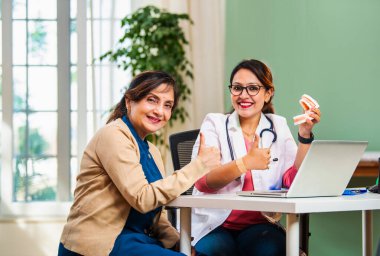 Hindistanlı genç bayan dişçi dişçi dişçi, dişçi mankeni kullanan yaşlı bir kadınla oral sağlığı tartışıyor, dişçi kliniğinde dizüstü bilgisayar, diş hijyeni, tedavi seçenekleri, tedavinin önemi...
