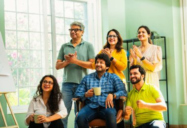 Modern girişim ofisindeki Hintli Asyalı genç şirket profesyonelleri, şık giyinmiş, el çırpıyor, başarıyı, başarıyı, takım çalışmasını, yeniliği, iş büyümesini kutluyorlar.