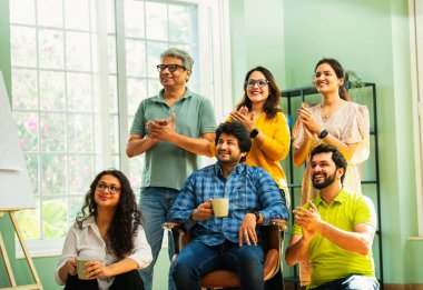 Modern girişim ofisindeki Hintli Asyalı genç şirket profesyonelleri, şık giyinmiş, el çırpıyor, başarıyı, başarıyı, takım çalışmasını, yeniliği, iş büyümesini kutluyorlar.