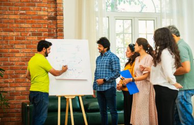 Hintli Asyalı genç girişimci gündelik kıyafetlerle iş stratejisini modern bir ofis, liderlik, takım çalışması konseptinde duran ekibine bir tabloya koyuyor.