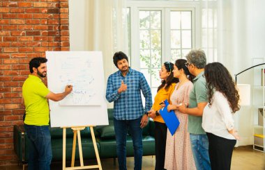 Hintli Asyalı genç girişimci gündelik kıyafetlerle iş stratejisini modern bir ofis, liderlik, takım çalışması konseptinde duran ekibine bir tabloya koyuyor.