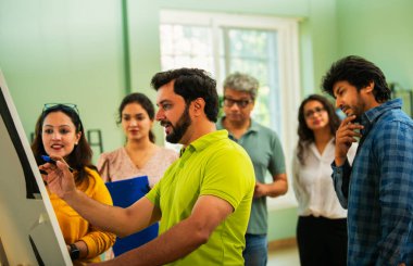 Hintli Asyalı genç girişimci gündelik kıyafetlerle iş stratejisini modern bir ofis, liderlik, takım çalışması konseptinde duran ekibine bir tabloya koyuyor.