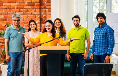 Hintli Asyalı genç girişimci konferansta hevesli iş adamlarına güvenerek konuşma yapıyor, mikrofonda podyumda dikiliyor ve ekibi arkadan alkışlıyor ve tezahürat yapıyor.