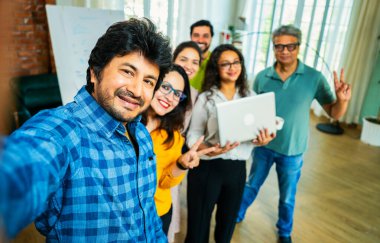 Hintli Asyalı genç girişimci gündelik kıyafetleriyle mutlu başlangıç ekibiyle birlikte modern bir ofiste takım çalışması, başarı ve kurumsal kültürü yakalarken selfie ya da fotoğraf çekiyor.