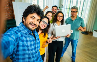 Hintli Asyalı genç girişimci gündelik kıyafetleriyle mutlu başlangıç ekibiyle birlikte modern bir ofiste takım çalışması, başarı ve kurumsal kültürü yakalarken selfie ya da fotoğraf çekiyor.
