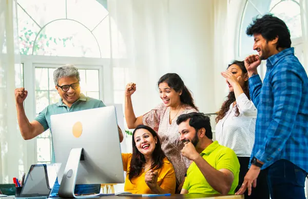 Hintli Asyalı genç iş adamları ya da teknoloji ortakları bilgisayarda çalışıyor, birden başarıyı beşlik çakarak kutluyorlar, modern rahat ofiste dizüstü bilgisayarla masada oturuyorlar.