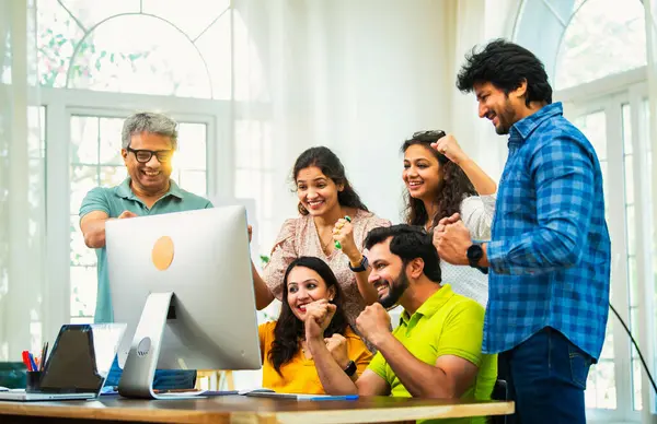 Hintli Asyalı genç iş adamları ya da teknoloji ortakları bilgisayarda çalışıyor, birden başarıyı beşlik çakarak kutluyorlar, modern rahat ofiste dizüstü bilgisayarla masada oturuyorlar.