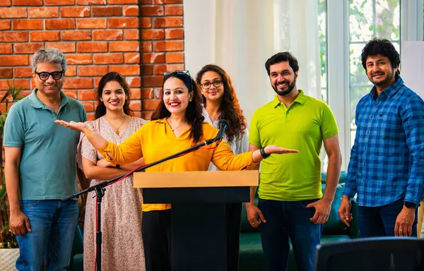 Hintli Asyalı genç girişimci konferansta hevesli iş adamlarına güvenerek konuşma yapıyor, mikrofonda podyumda dikiliyor ve ekibi arkadan alkışlıyor ve tezahürat yapıyor.