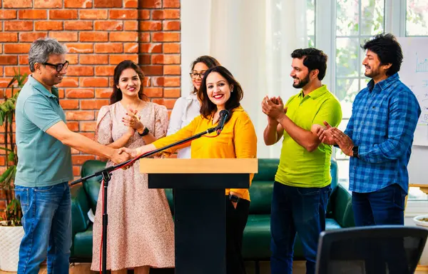 Hintli Asyalı genç girişimci konferansta hevesli iş adamlarına güvenerek konuşma yapıyor, mikrofonda podyumda dikiliyor ve ekibi arkadan alkışlıyor ve tezahürat yapıyor.