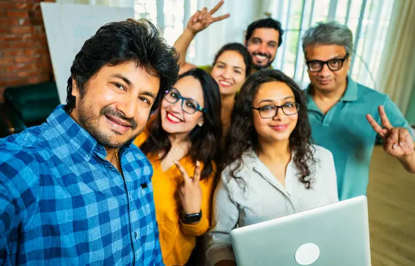 Hintli Asyalı genç girişimci gündelik kıyafetleriyle mutlu başlangıç ekibiyle birlikte modern bir ofiste takım çalışması, başarı ve kurumsal kültürü yakalarken selfie ya da fotoğraf çekiyor.