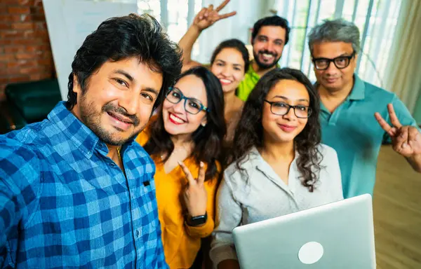 Hintli Asyalı genç girişimci gündelik kıyafetleriyle mutlu başlangıç ekibiyle birlikte modern bir ofiste takım çalışması, başarı ve kurumsal kültürü yakalarken selfie ya da fotoğraf çekiyor.