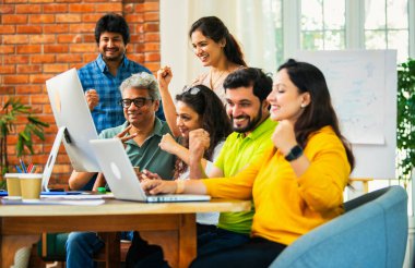 Hintli Asyalı genç iş adamları ya da teknoloji firması ortakları bilgisayarda çalışıyor, birden başarıyı beşlik çakarak kutluyorlar, modern rahat ofiste masa başında oturuyorlar.