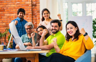 Hintli Asyalı genç iş adamları ya da teknoloji firması ortakları bilgisayarda çalışıyor, birden başarıyı beşlik çakarak kutluyorlar, modern rahat ofiste masa başında oturuyorlar.