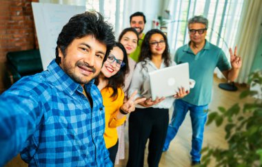 Hintli Asyalı genç girişimci gündelik kıyafetleriyle mutlu başlangıç ekibiyle birlikte modern bir ofiste takım çalışması, başarı ve kurumsal kültürü yakalarken selfie ya da fotoğraf çekiyor.