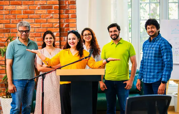 Hintli Asyalı genç girişimci konferansta hevesli iş adamlarına güvenerek konuşma yapıyor, mikrofonda podyumda dikiliyor ve ekibi arkadan alkışlıyor ve tezahürat yapıyor.