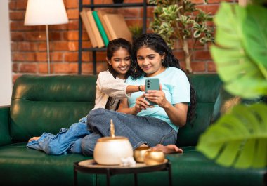 Hintli Asyalı genç anne kız evdeki rahat modern koltukta oturuyor, akıllı telefonu online alışveriş için kullanıyor, interneti tarıyor, sosyal medyayı karıştırıyor, video görüşmeleri yapıyor, dijital yaşam tarzının tadını çıkarıyor.