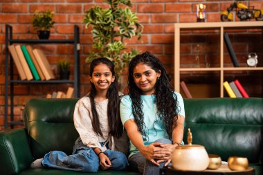 Hintli Asyalı genç anne ve tatlı küçük kızının portresi sıcak bir şekilde gülümsüyor ve doğrudan kameraya bakıyor rahat bir kanepede otururken şık, müsrif modern ev içinde.