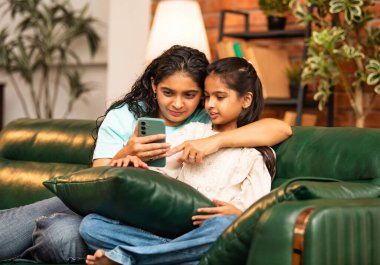 Hintli Asyalı genç anne kız evdeki rahat modern koltukta oturuyor, akıllı telefonu online alışveriş için kullanıyor, interneti tarıyor, sosyal medyayı karıştırıyor, video görüşmeleri yapıyor, dijital yaşam tarzının tadını çıkarıyor.