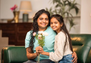 Hintli Asyalı genç anne, küçük kızına kanepede sarılıp, kalpten bir Anneler Günü 'nü kutlarken, kızının yaptığı çiçekler ve hediyelere bakarken aile bağlarını sergiliyor, sevgisini sergiliyor.