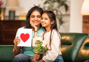 Hintli Asyalı genç anne, küçük kızına kanepede sarılıp, kalpten bir Anneler Günü 'nü kutlarken, kızının yaptığı çiçekler ve hediyelere bakarken aile bağlarını sergiliyor, sevgisini sergiliyor.