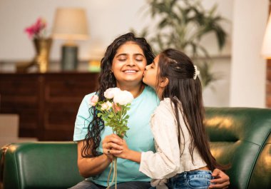 Hintli Asyalı genç anne, küçük kızına kanepede sarılıp, kalpten bir Anneler Günü 'nü kutlarken, kızının yaptığı çiçekler ve hediyelere bakarken aile bağlarını sergiliyor, sevgisini sergiliyor.